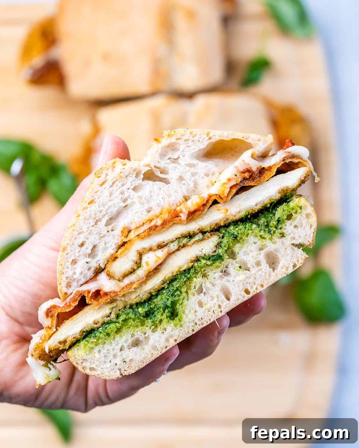 Crispy Chicken Parmesan Sandwich Outshines Takeout 7 A hand holding a perfectly assembled Chicken Parmesan Sandwich, showing off its crispy breaded chicken, melted mozzarella, vibrant red marinara sauce, and bright green pesto on toasted ciabatta bread. The background is softly blurred, highlighting the sandwich as the main focus.