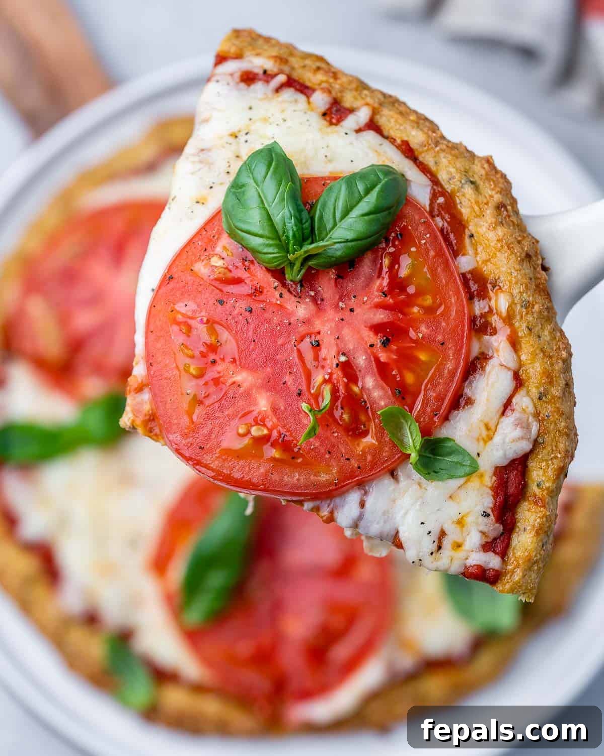 A perfectly cooked slice of chicken crust pizza, topped with melted cheese, tomato, and basil, being held up on a spatula.