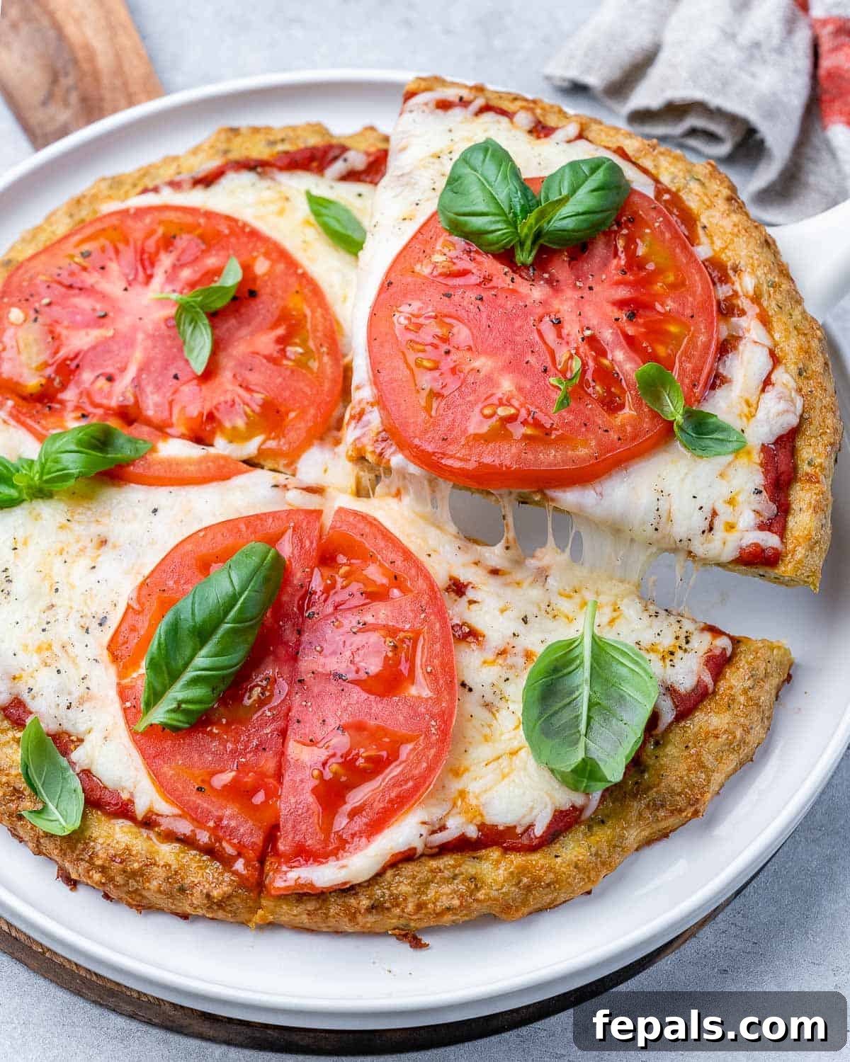 A close-up shot of a chicken crust pizza slice on a white plate, topped with vibrant tomato slices and fresh basil, showcasing its golden crust.