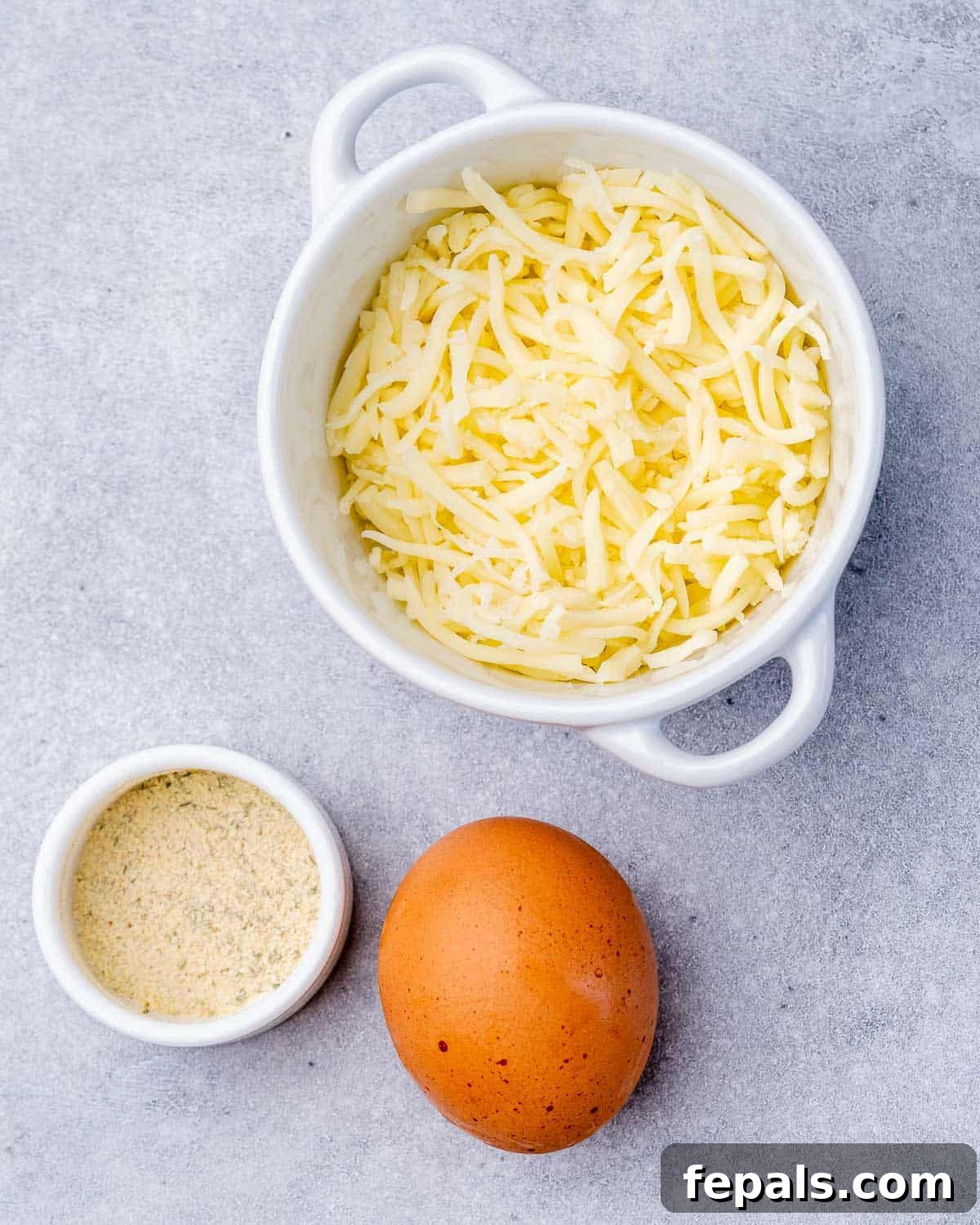 Overhead view of a white bowl filled with shredded mozzarella, a small ramekin of garlic salt, and one whole egg.