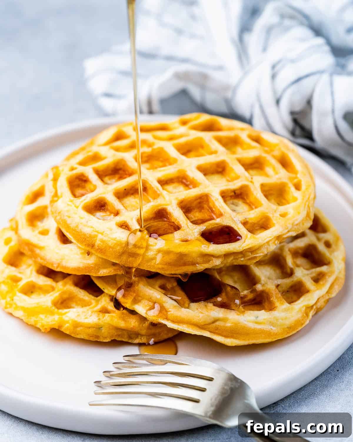 Three warm chaffles stacked on a white plate, with syrup being poured over the top.