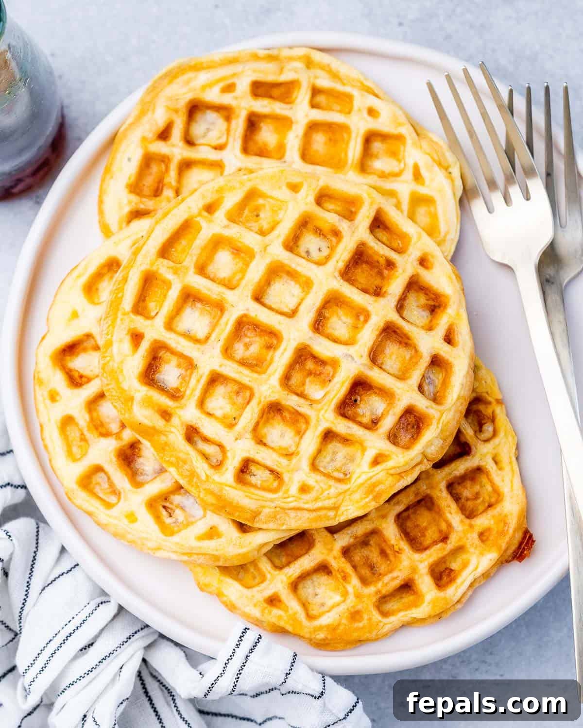 A full plate of freshly cooked chaffles next to two silver forks and a striped cloth napkin.