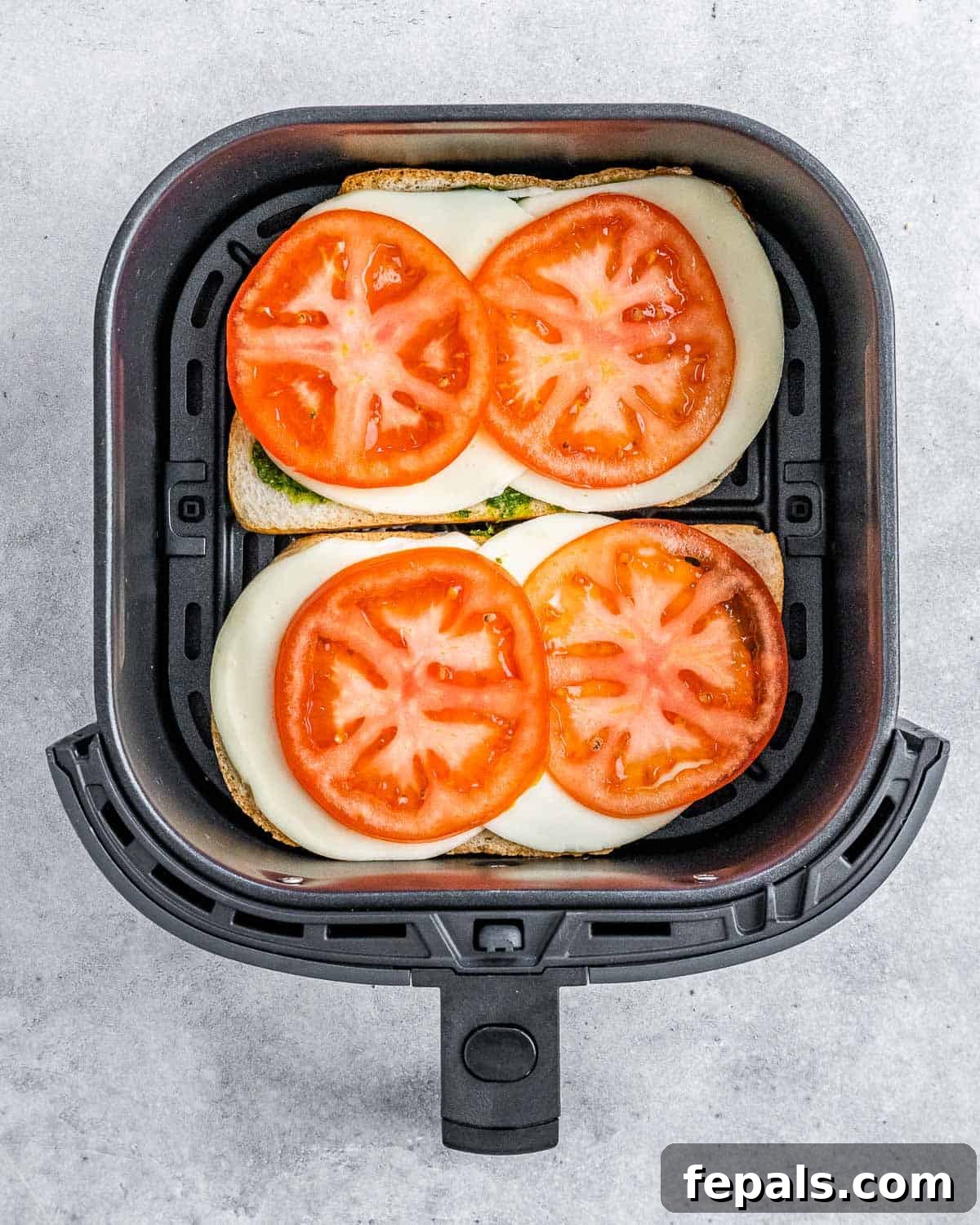 Two slices of sandwich bread in an air fryer basket, topped with mozzarella cheese and thin slices of fresh tomato.