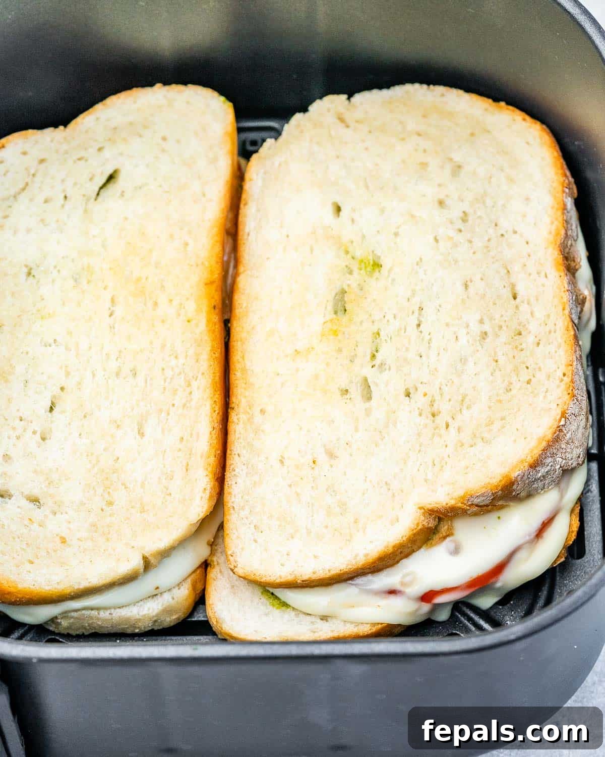A partially cooked cheese and tomato sandwich inside an air fryer basket, showing the melting cheese and golden bread.