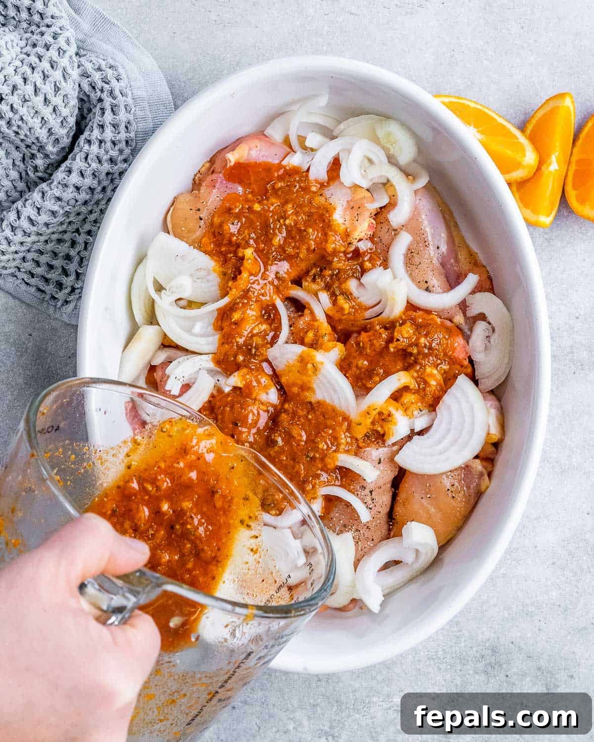 Crispy Pulled Chicken Carnitas 4 Pouring a vibrant orange-garlic marinade over boneless chicken thighs and thinly sliced onions in a deep baking dish.