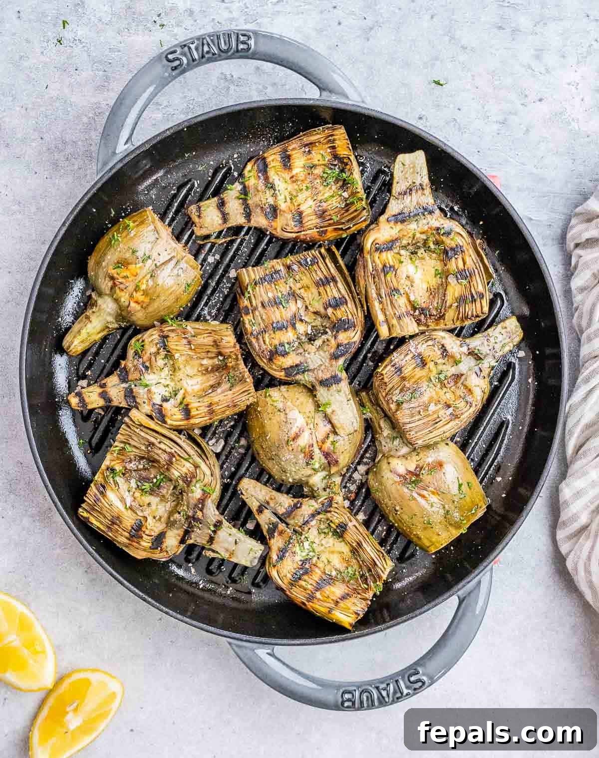 Smoky Grilled Artichoke Halves 2 Two halves of grilled artichokes, perfectly charred, garnished with fresh lemon wedges and herbs, presented on a rustic grill pan.