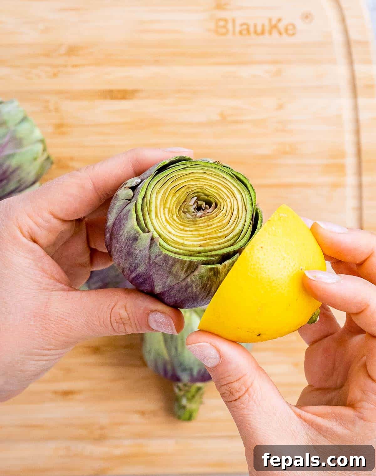 Smoky Grilled Artichoke Halves 5 A hand gently rubs a lemon half over the freshly cut surface of an artichoke to prevent browning and add zest.