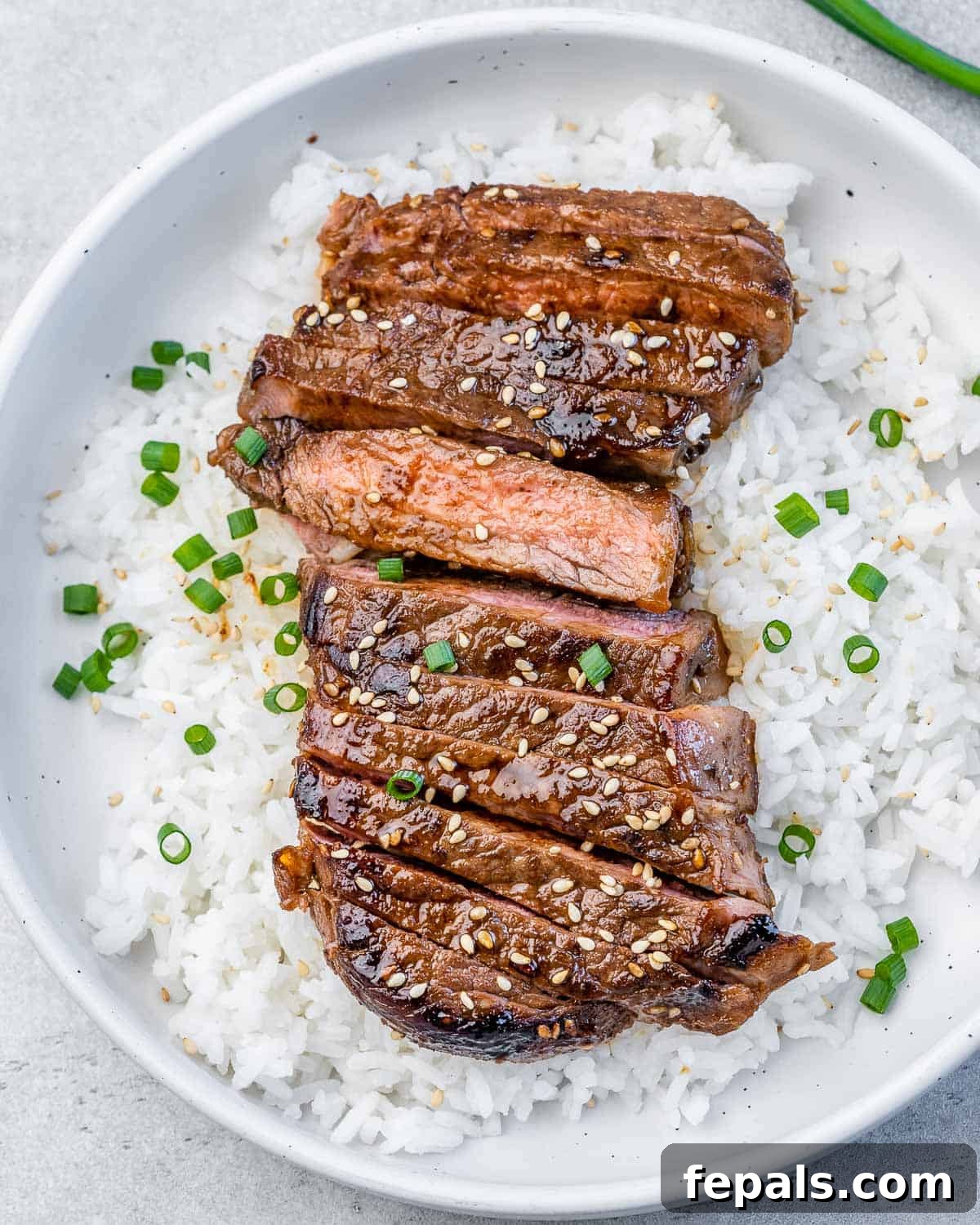 Sweet and Savory Teriyaki Steak 2 A round white plate with white rice topped with sliced teriyaki steak garnished with chopped green onions and sesame seeds.