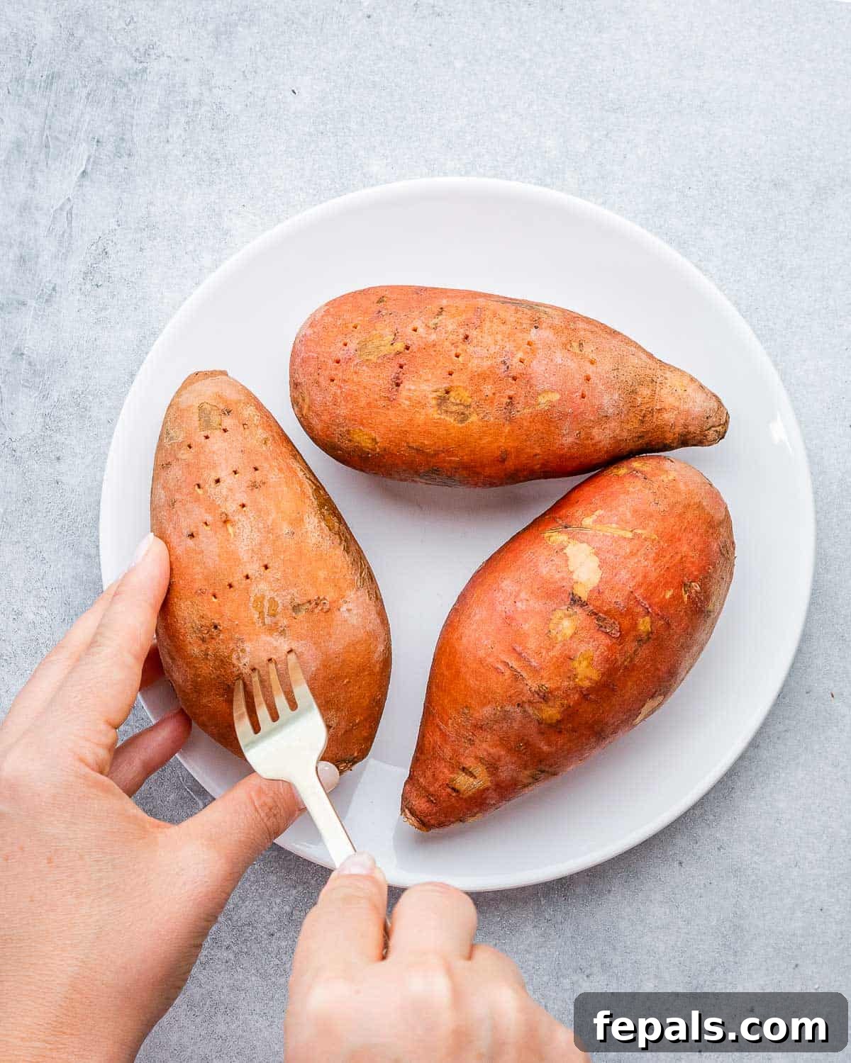 A fork piercing several sweet potatoes on a plate, preparing them for cooking.