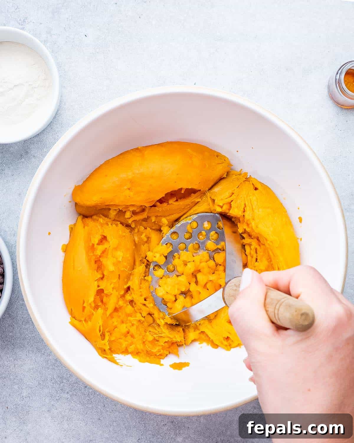 Cooked sweet potatoes being mashed into a smooth puree in a large bowl with a potato masher.