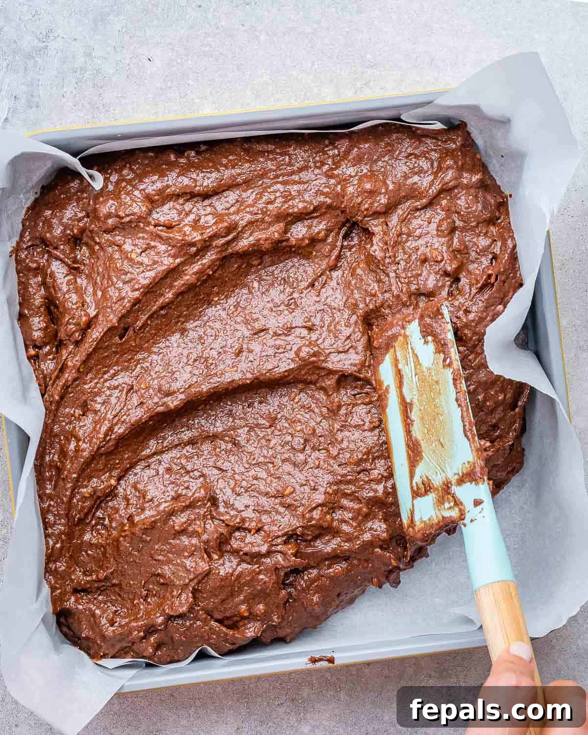 Smooth brownie batter being evenly spread into a square baking pan lined with parchment paper, preparing it for baking.