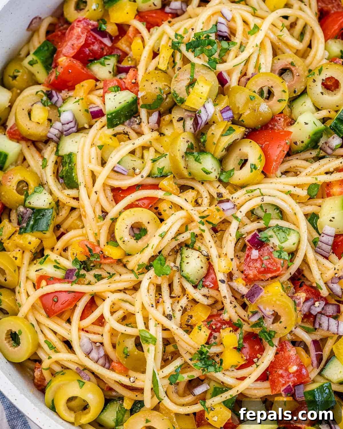 Refreshing Spaghetti Salad 9 A close-up of a delightful spaghetti salad in a white ceramic dish, featuring long strands of pasta beautifully intertwined with colorful diced vegetables and an abundance of sliced green olives, ready to be enjoyed.