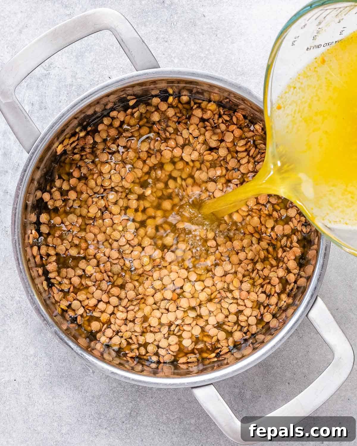 Pouring vegetable broth into a pot with rinsed green lentils, ready for cooking.