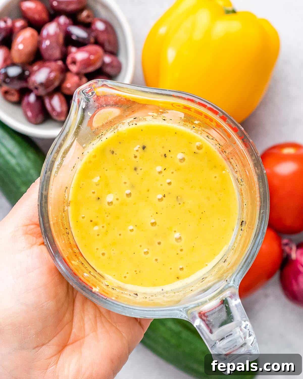 A hand holding a Pyrex measuring cup filled with freshly whisked lemon-Dijon dressing, showcasing its vibrant yellow color and smooth texture.