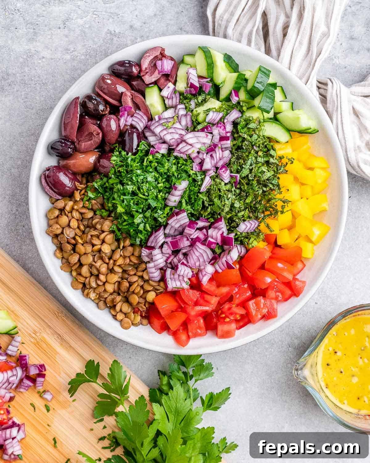 Cooked green lentils and colorful chopped vegetables in a white bowl, ready to be drizzled with the lemon-Dijon dressing shown nearby in a glass.