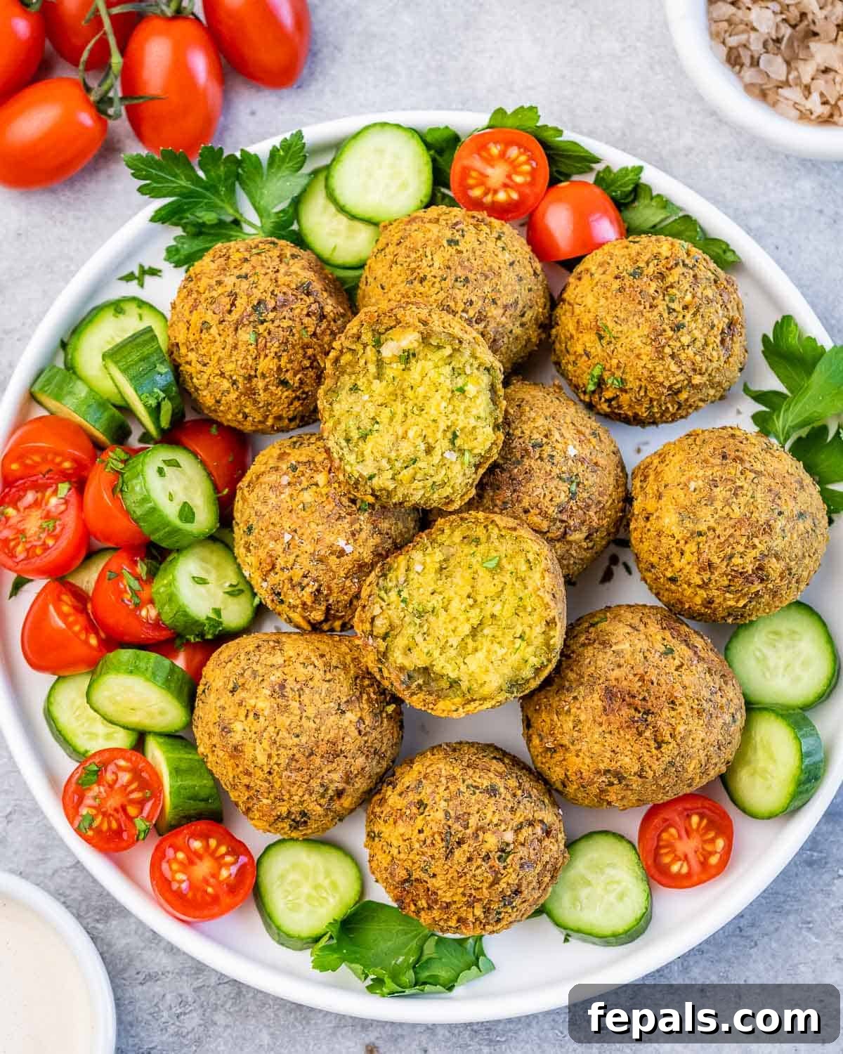 Crispy Homemade Falafel Made Simple 2 Top view of a plate of crispy falafel balls surrounded by sliced cucumbers, cherry tomatoes, and parsley on a light background.