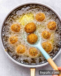 Falafel balls frying in hot oil inside a deep skillet with a slotted spoon lifting one out.