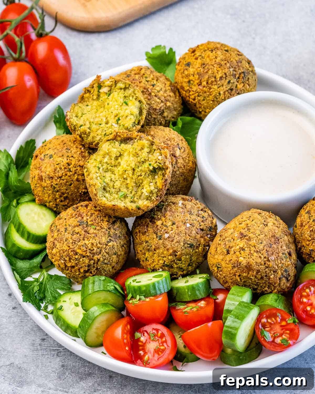 Crispy Homemade Falafel Made Simple 14 Falafel balls served with cucumber, tomato, parsley, and a creamy dipping sauce on a round white plate.