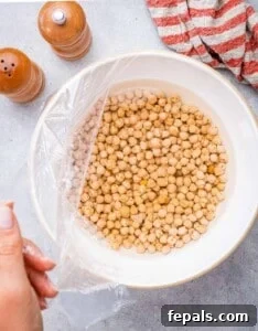 Bowl of dried chickpeas soaking in water, covered with plastic wrap.