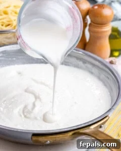 Thick cottage cheese Alfredo sauce being poured from a measuring cup into a skillet with sautéed garlic.
