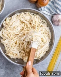 Hand stirring cooked spaghetti in a pan to evenly combine with the Alfredo sauce.