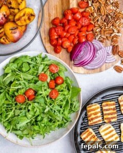 Arugula and cherry tomatoes in a bowl beside sliced red onions, toasted pecans, grilled peaches, and halloumi on a cutting board.