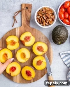 Halved peaches with pits removed on a cutting board, next to cherry tomatoes, pecans, and an avocado.
