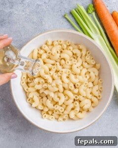Aloha Macaroni Salad 11 Apple cider vinegar being poured over freshly cooked elbow macaroni in a white bowl.