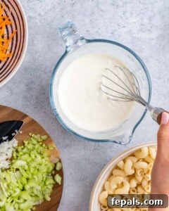 Aloha Macaroni Salad 12 Hand whisking together mayonnaise and milk in a glass measuring cup, with chopped celery, onion, and cooked macaroni nearby.