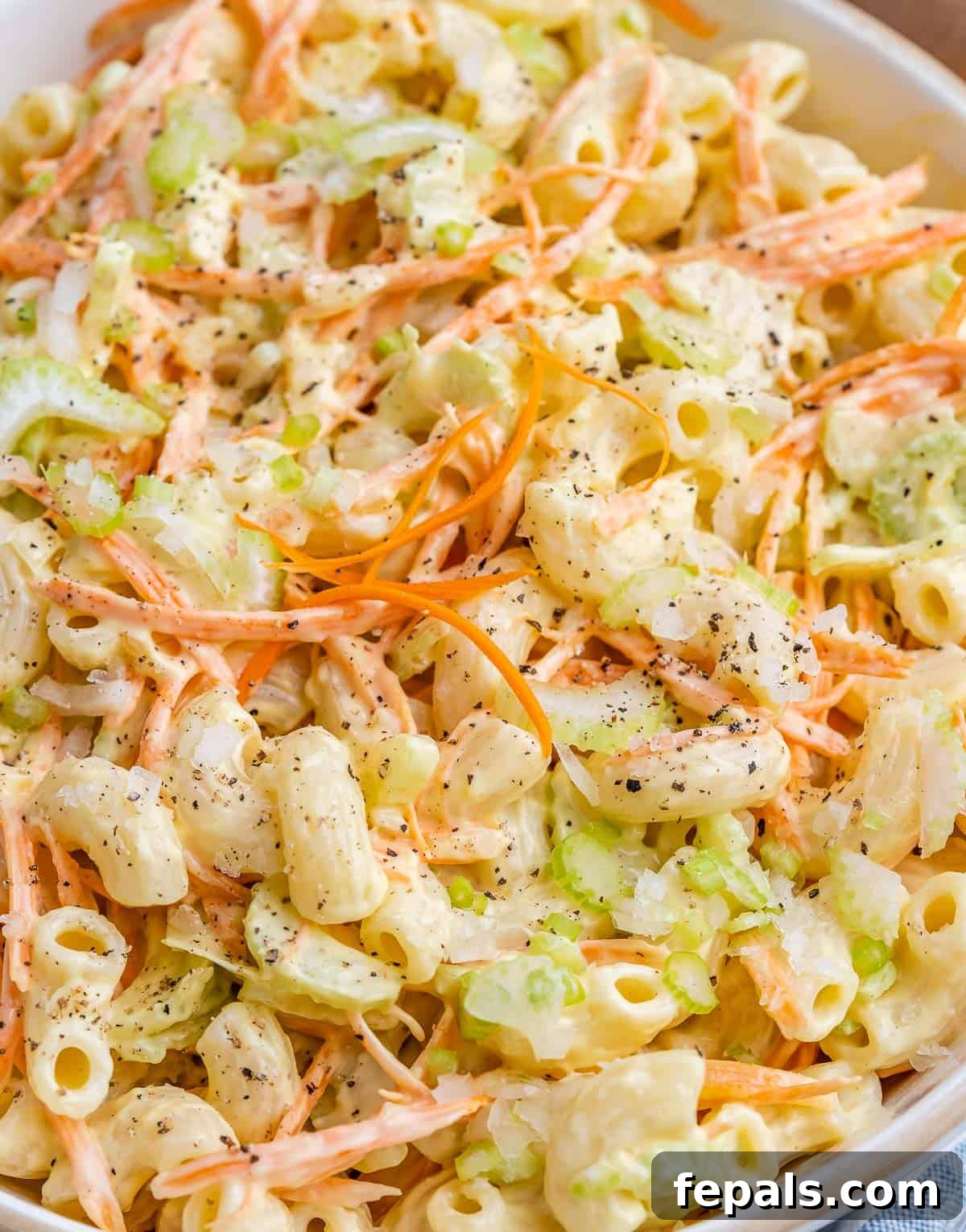 Aloha Macaroni Salad 3 A close-up view of the irresistible Hawaiian mac salad, showcasing its creamy pasta intertwined with bright orange shredded carrots, crisp green celery pieces, and a generous sprinkle of black pepper, highlighting its appealing texture and freshness.