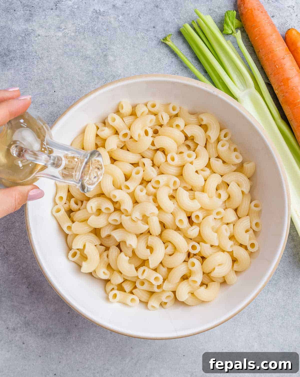 Aloha Macaroni Salad 5 Freshly cooked elbow macaroni being gently drizzled with apple cider vinegar in a pristine white bowl, ensuring each piece is coated for optimal flavor absorption.