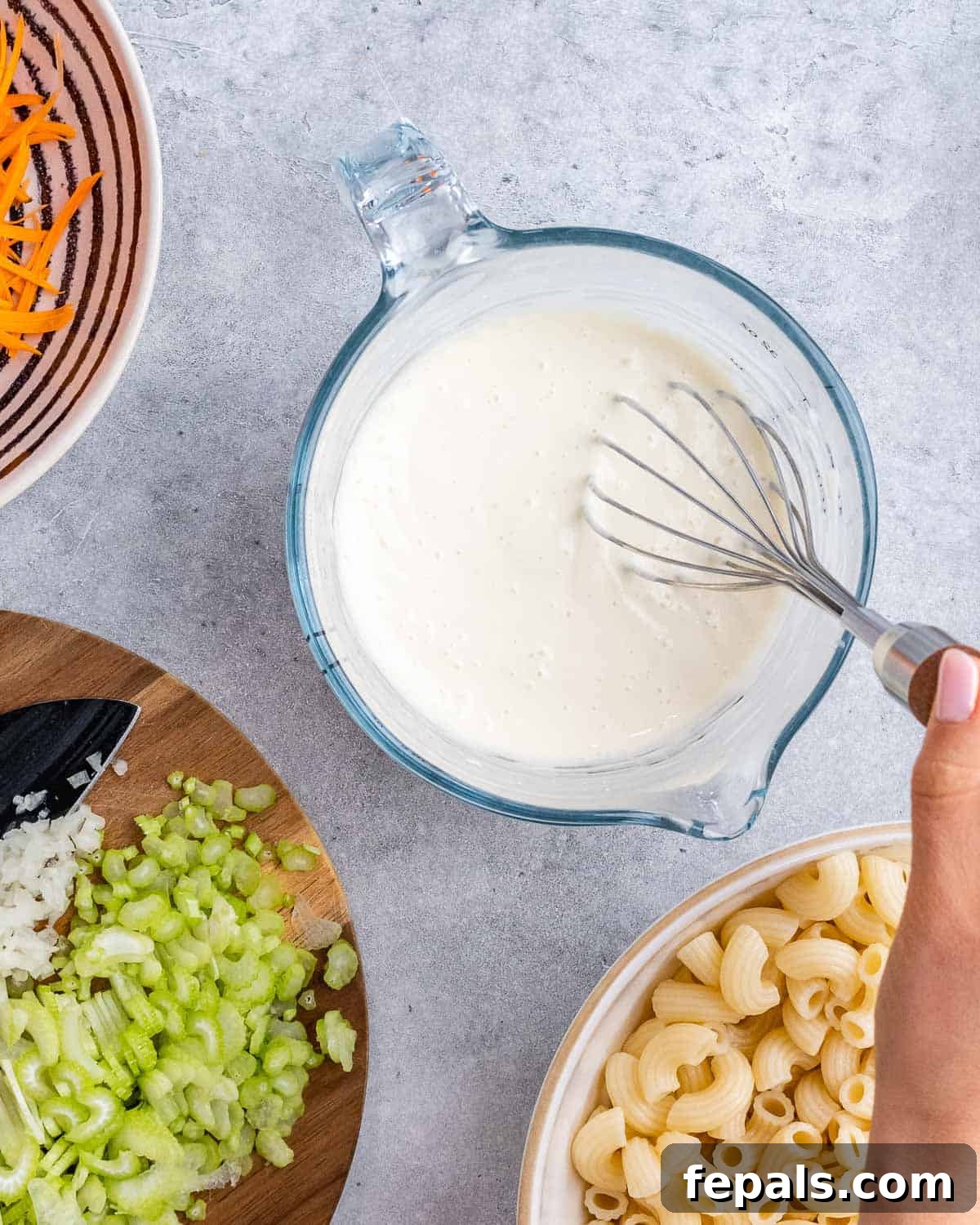 Aloha Macaroni Salad 6 A hand expertly whisking together creamy mayonnaise and a splash of milk in a clear glass measuring cup, with prepped ingredients like chopped celery, grated onion, and cooked macaroni visible nearby, illustrating the dressing preparation.