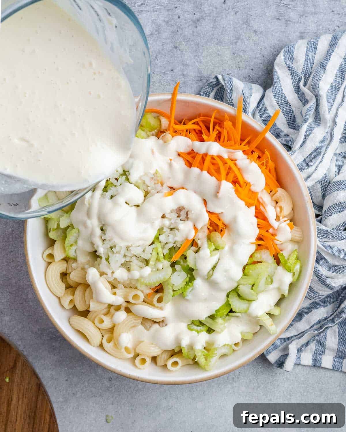 Aloha Macaroni Salad 8 The rich, creamy mayo and milk dressing gracefully pouring over the vibrant mix of chopped vegetables and perfectly cooked macaroni in a spacious mixing bowl, initiating the final stage of salad preparation.