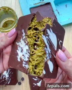 A hand breaking a solidified chocolate bar, revealing the green pistachio filling.