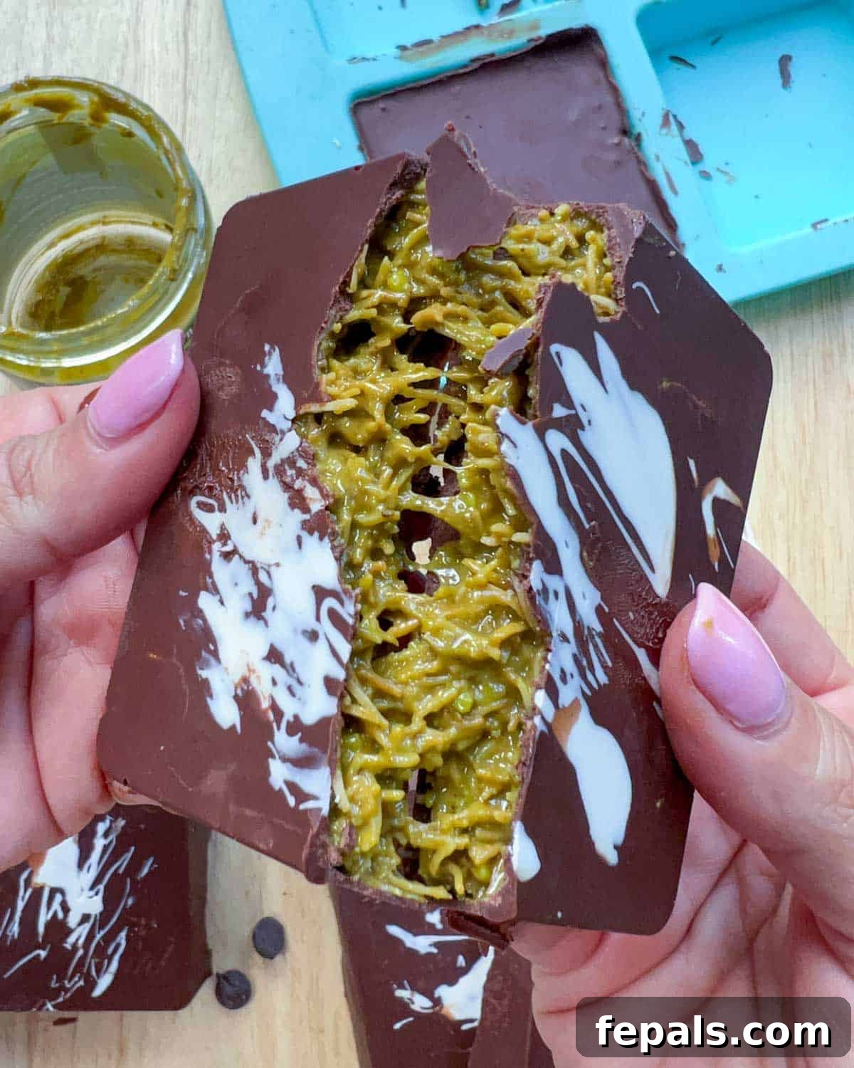A hand breaking a bar of chocolate, revealing the rich green pistachio filling and crunchy texture inside.