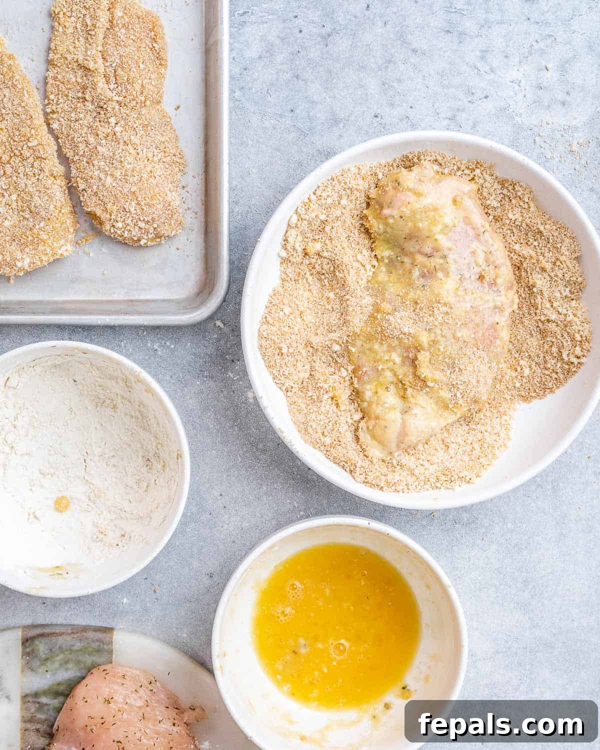Ultimate Chicken Parm Bake 5 Dredging station with chicken cutlets coated in flour, egg, and breadcrumbs.