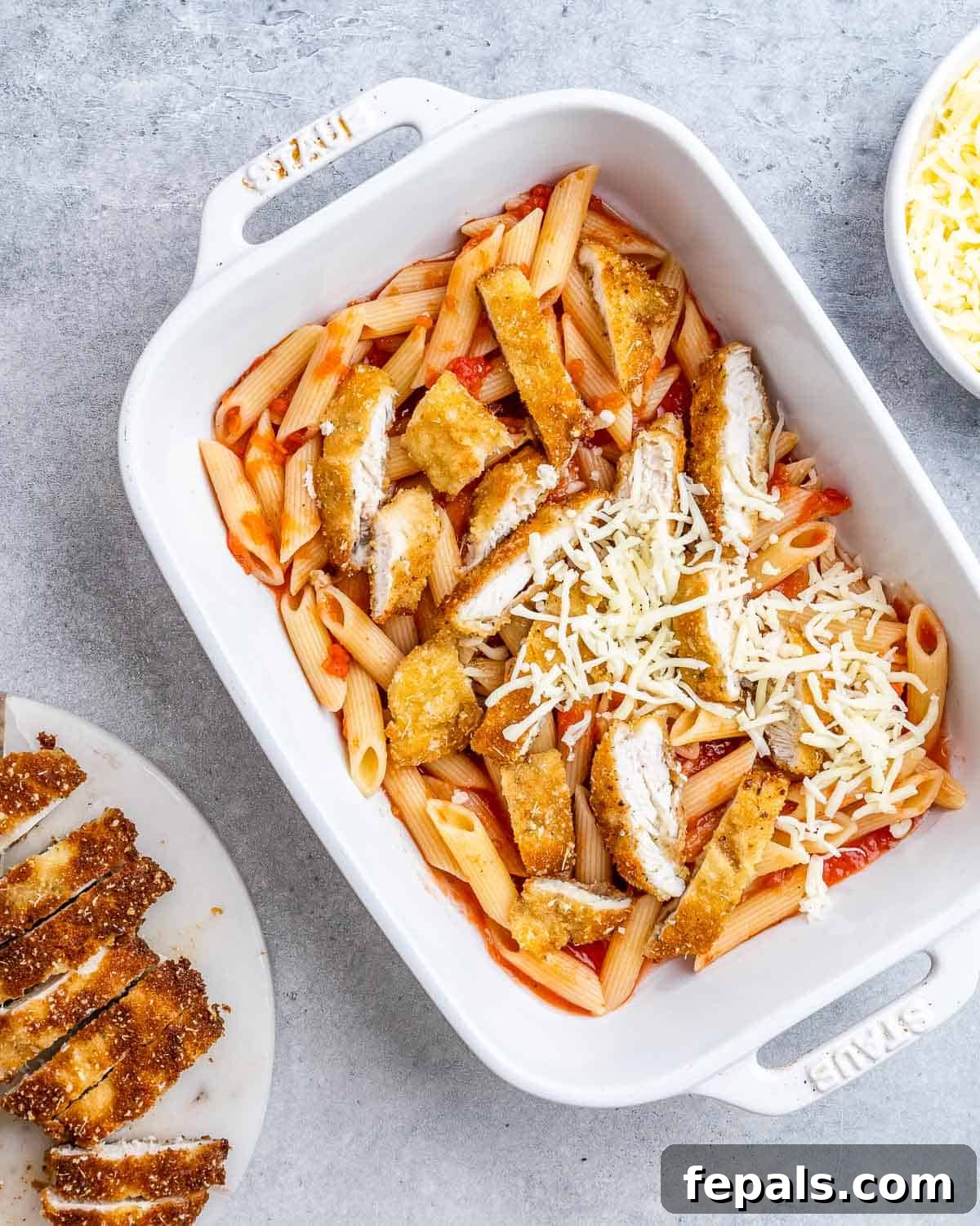 Ultimate Chicken Parm Bake 7 Sprinkling mozzarella cheese over the top of a layered casserole with chicken and pasta.