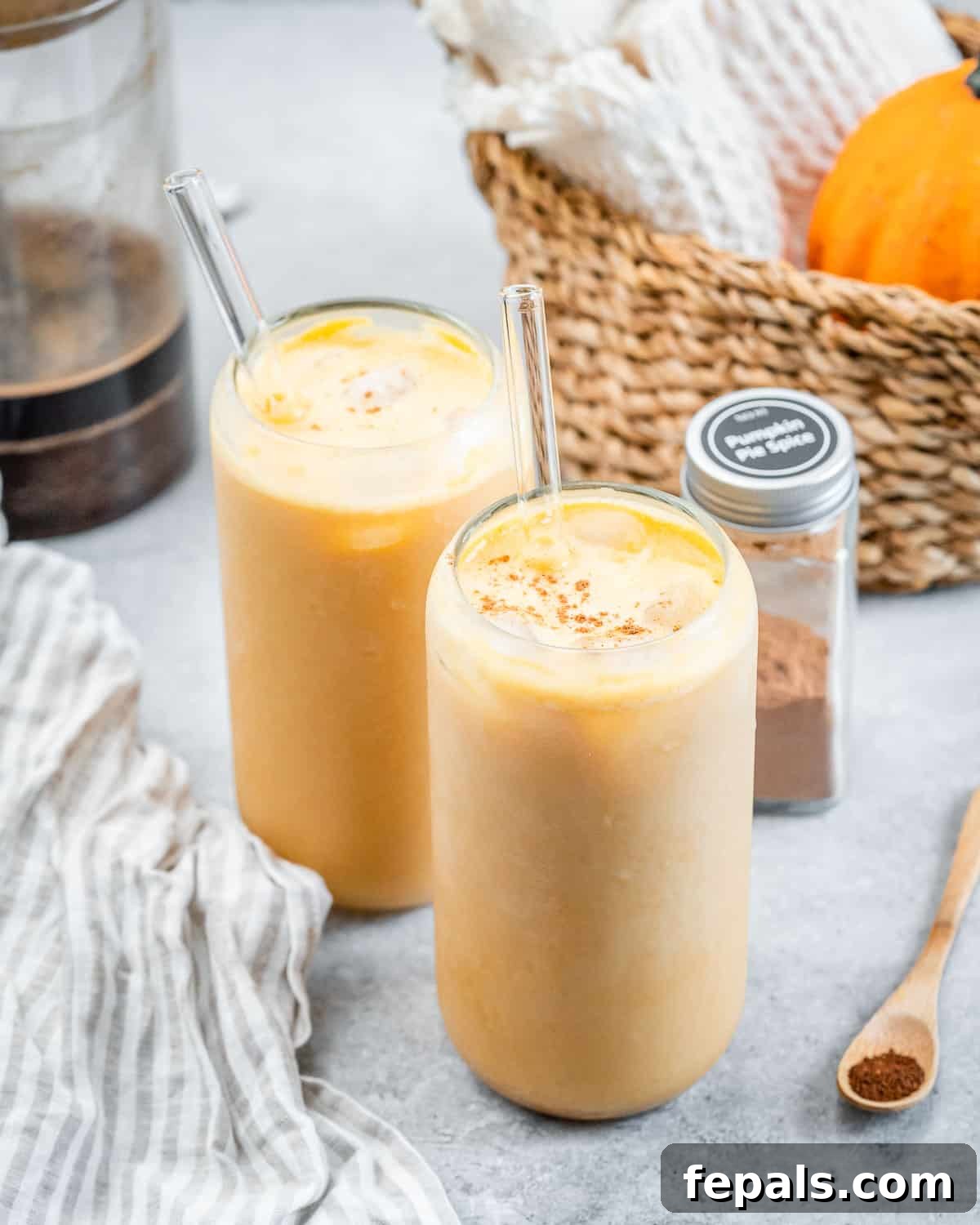 Two glasses of homemade Pumpkin Cream Cold Brew, garnished with cinnamon sticks and a sprinkle of pumpkin pie spice, featuring clear glass straws, set against a warm autumn-themed background.