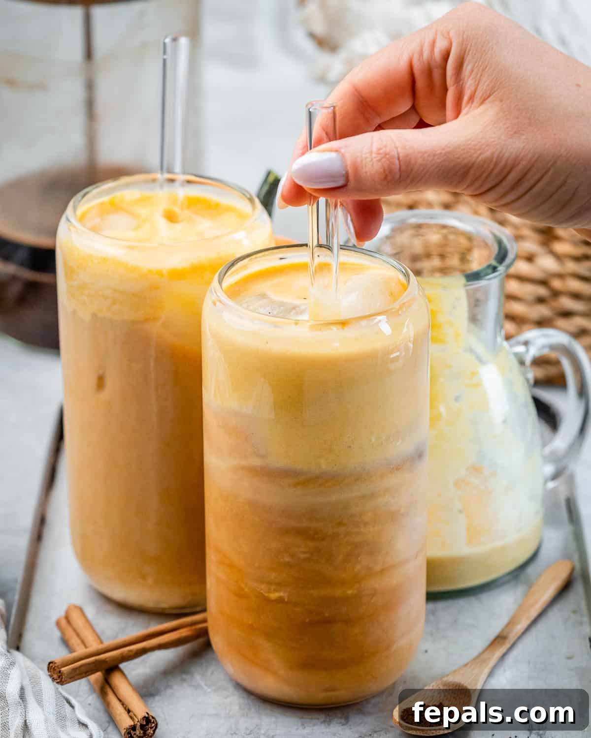 A woman's hand gently stirring a Pumpkin Cream Cold Brew in a tall glass with a glass straw, highlighting the smooth texture and appealing layers of the drink.