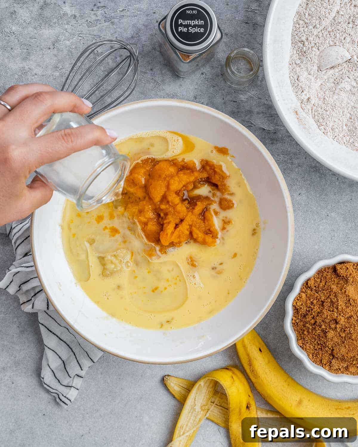 Pouring a liquid ingredient, possibly melted coconut oil, into a bowl containing a mixture of pumpkin puree, eggs, and vanilla extract, with a whisk nearby.