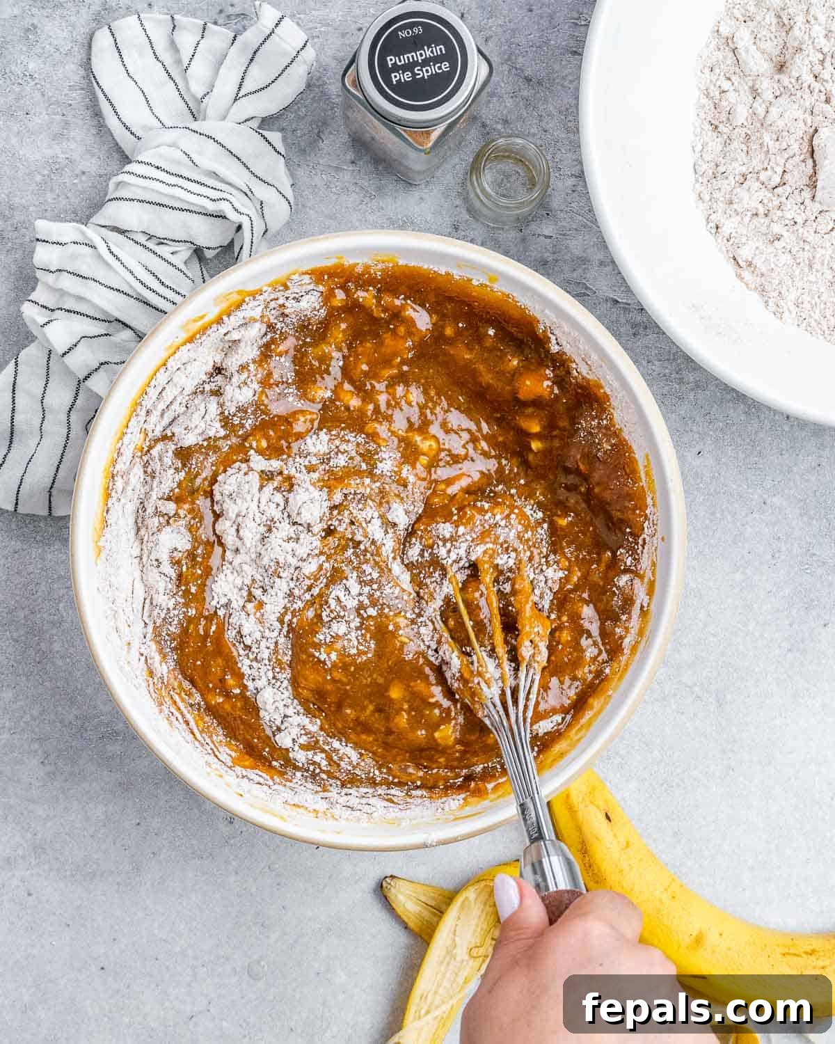 Gently stirring the dry flour and spice mixture into the wet pumpkin and banana ingredients in a large white bowl, ensuring no lumps remain.