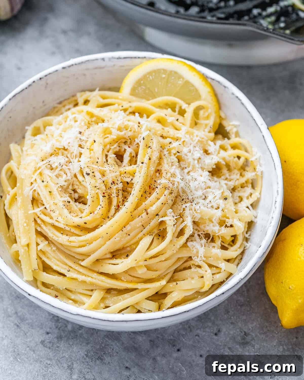 Close up of Pasta al Limone in a white bowl, surrounded by fresh lemons, garnished with Parmesan.