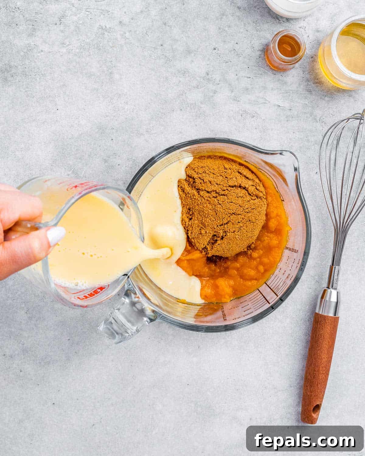 Wholesome Pumpkin Pie 5 A hand pouring milk into a large mixing bowl containing a creamy mixture of pumpkin puree, eggs, coconut sugar, honey, and spices.
