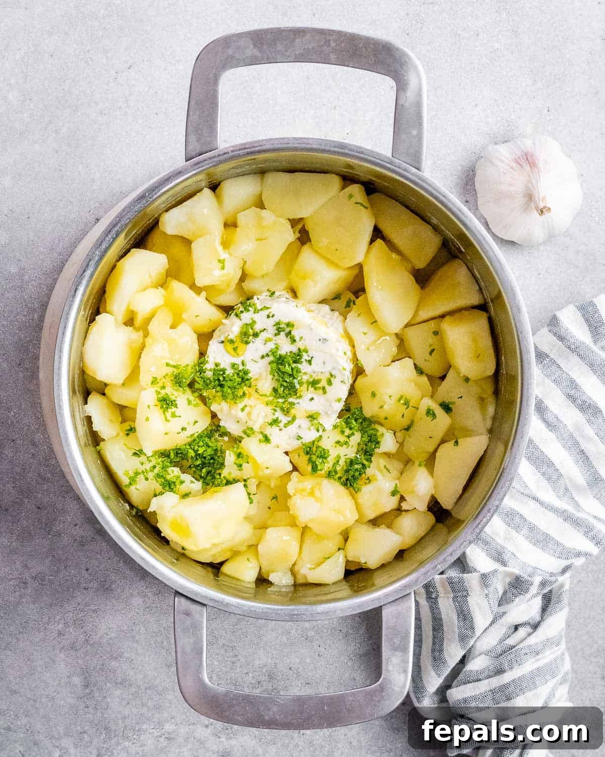 A sprinkle of fresh parsley, chunks of Boursin cheese, and a pour of olive oil being added to hot, drained potatoes in a pot.