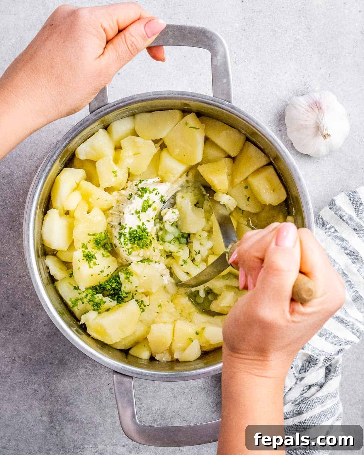 A hand mashing potatoes in a pot with a sturdy, silver potato masher, showing the early stages of combining ingredients.