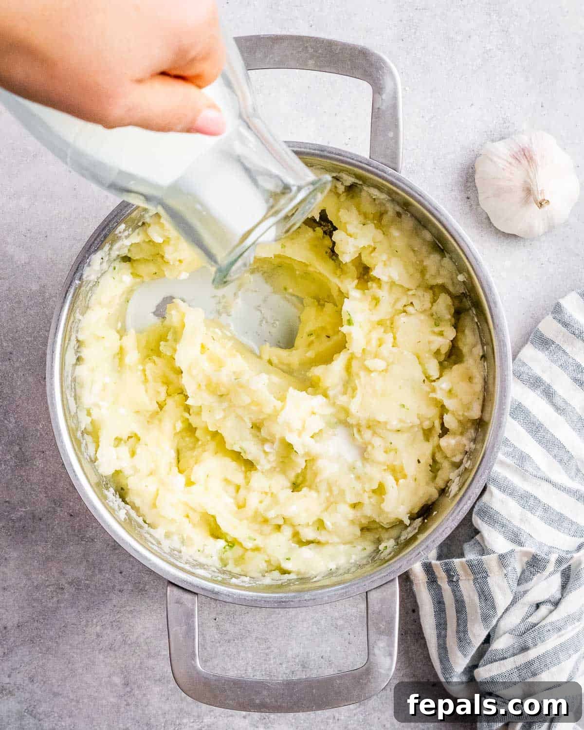Warm milk being slowly poured into a pot of mashed potatoes as they are gently stirred to achieve the desired consistency.