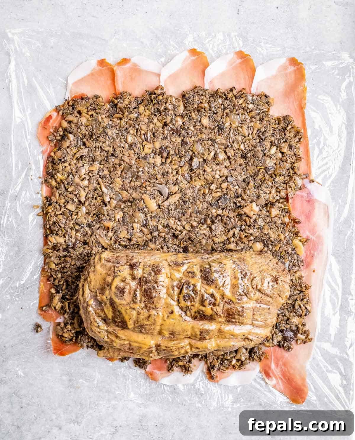Regal Beef Wellington 6 The beef tenderloin, coated in mushroom duxelles and prosciutto, is being tightly rolled in cling film.