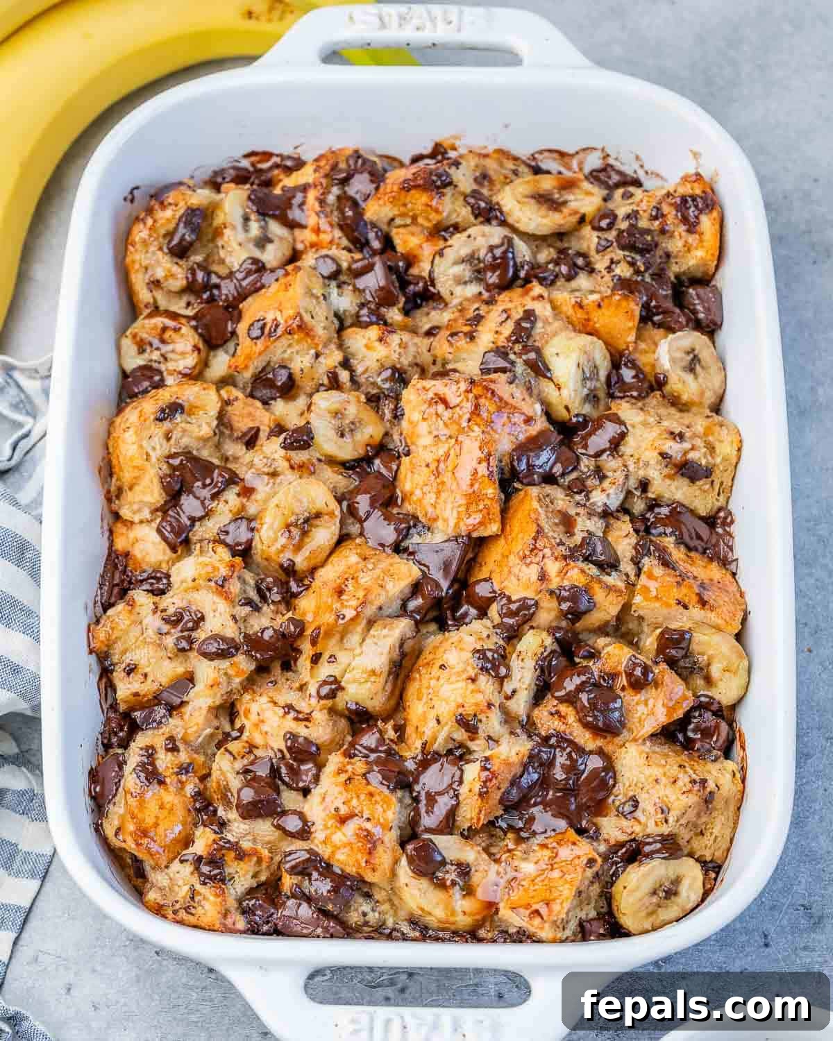 A beautifully baked banana bread pudding casserole, golden brown and topped with melted chocolate chips and fresh banana slices, served in a rustic white baking dish. The pudding is rich and inviting.