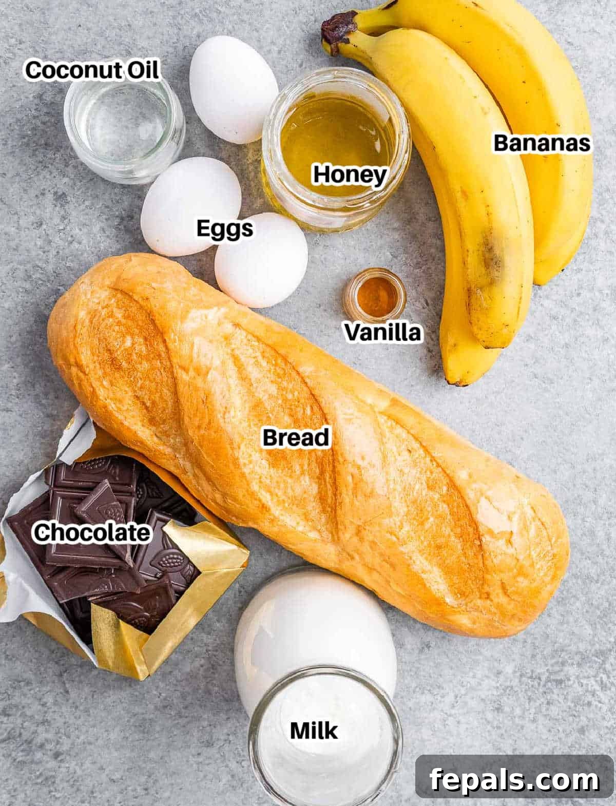A collection of fresh ingredients laid out neatly for making banana bread pudding. Includes ripe bananas, eggs, milk, honey, vanilla extract, cubed bread, and chopped dark chocolate.