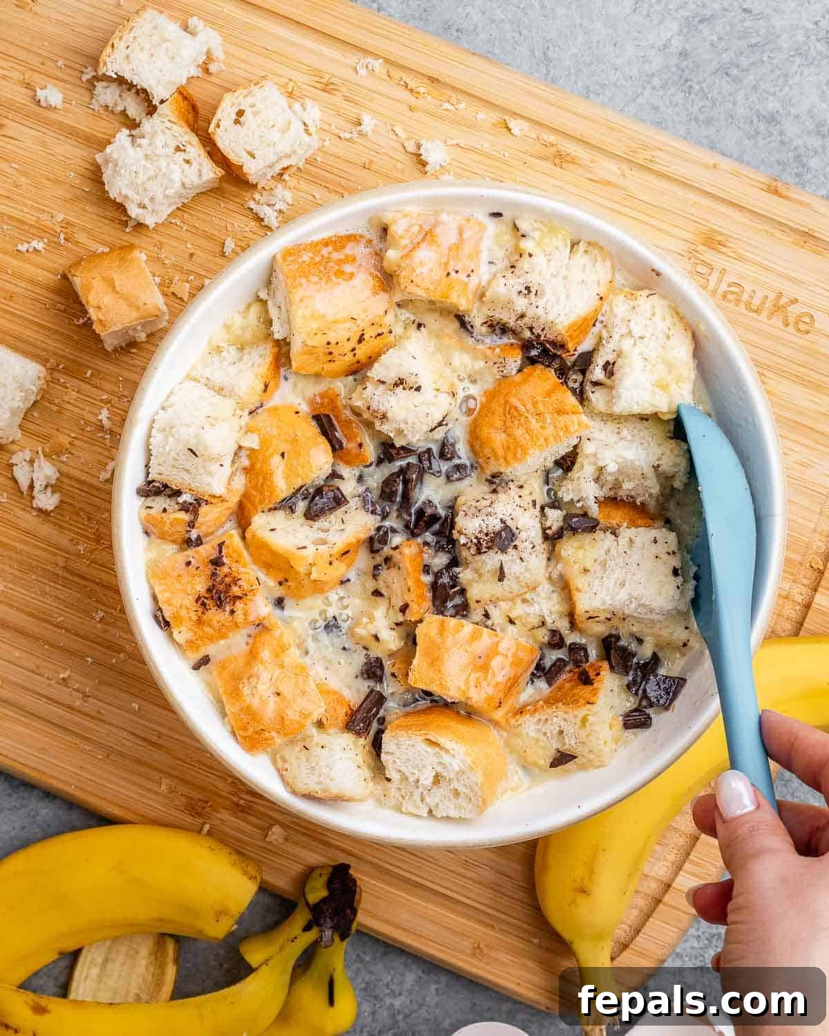 Chopped bread cubes gently folded into the banana and egg mixture in a large bowl, along with chocolate pieces, preparing the pudding.